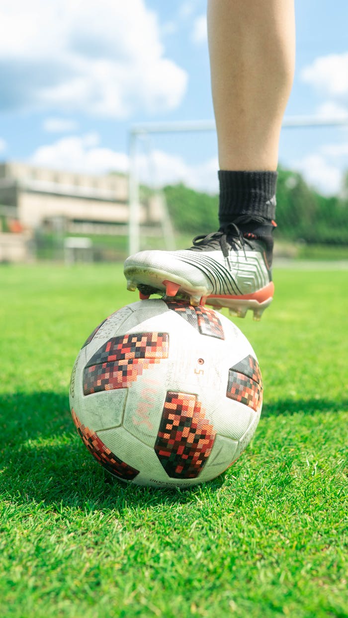 pexels-photo-13422993 Close-up of a soccer ball with a player's foot on it, set on a vibrant green soccer field during a sunny day.