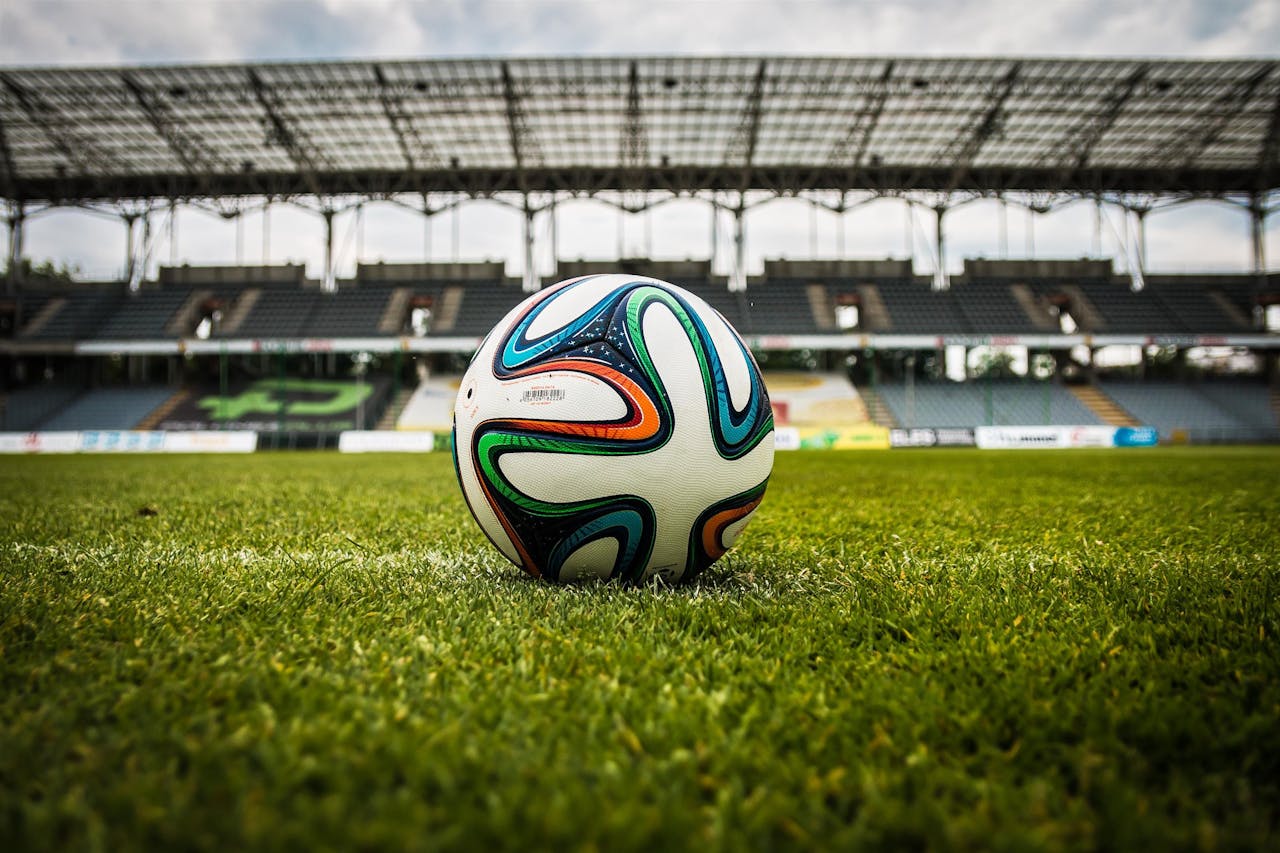 the-ball-stadion-football-the-pitch-47730 A vibrant soccer ball rests on a lush green field inside an empty stadium, capturing the essence of sports.